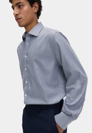 Ung mand modeller en slim-fit, langærmet, blå og hvid ternet skjorte, stukket ned i mørkeblå bukser.