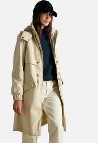 Sieviete, kas valkā bēšas krāsas lietusmantu ar kapuci, tumši zaļu un tumši zilu argyle adījumu, krēmkrāsas bikses un melnu beisbola cepuri.