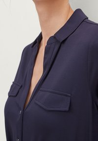 Blouse bleu marine avec un col, dotée de deux poches poitrine et d'une fermeture à boutons. Tissu lisse avec une texture nette.