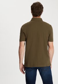 Zielona oliwkowa koszulka polo z krótkim rękawem i kołnierzykiem, o gładkiej fakturze i bocznych rozcięciach, noszona z niebieskimi dżinsami.