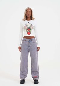 Frau mit blonden Haaren, die ein weißes langärmliges Crop-Top mit floralem Herzdesign, lockere graue Jeans und schwarze Plateauschuhe trägt.