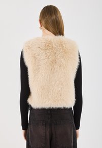 Faux fur väst i beige med en mjuk och fluffig textur. Har en åtsittande design och bärs över en svart ribbad långärmad tröja och mörka byxor.