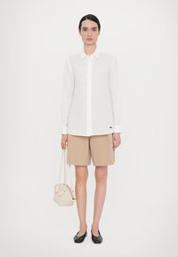 White long-sleeve shirt, button-up with collar, paired with beige shorts. Holding a woven beige bag, wearing black slip-on shoes.