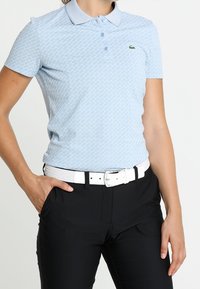 Ljusblå kortärmad pikétröja med ett strukturerat diamantmönster, knappslå och broderad grön logotyp på bröstet, kombinerad med svarta byxor och ett vitt bälte.