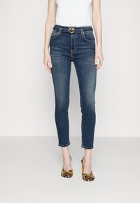 Jeans ajustados de talle alto en color azul oscuro, con un cinturón decorativo de hebilla dorada, combinados con una blusa blanca y tacones de serpiente.