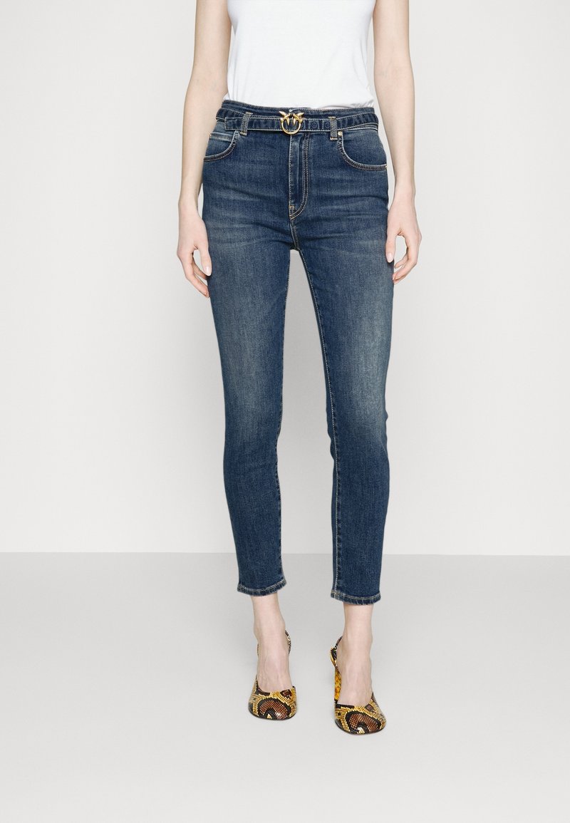 Jeans ajustados de talle alto en color azul oscuro, con un cinturón decorativo de hebilla dorada, combinados con una blusa blanca y tacones de serpiente.