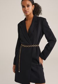 Rochie bleumarin din blazer cu decolteu adânc în V, croială ajustată și mâneci lungi. Prezintă un brâu din lanț auriu cu linkuri octogonale și un accent atârnat.