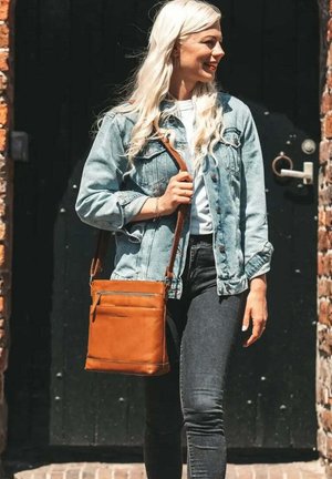 Tan leather crossbody bag with a front zip pocket, smooth texture, and adjustable strap. Worn with a light denim jacket and dark jeans.