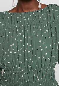 Vestido de tela verde con escote fruncido y cintura elástica, con un patrón floral en blanco y acentos en negro. Textura suave.