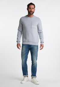 Felpa grigia con tasca sul petto e testo stampato, abbinata a jeans blu e sneakers bianche. Presenta un collo tondo e maniche lunghe.