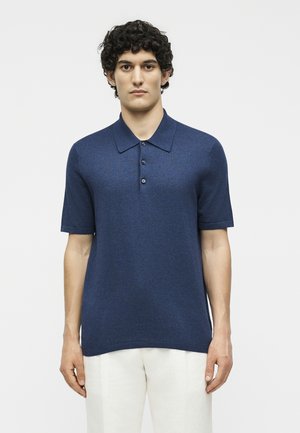 Άνδρας με σγουρά σκούρα μαλλιά φοράει μπλε σκούρο polo με κοντά μανίκια και λευκό παντελόνι, στέκεται μπροστά σε απλό λευκό φόντο.