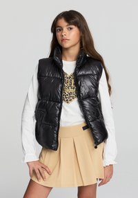 Giubbotto imbottito nero con colletto alto e chiusura con zipper, abbinato a una camicia bianca a maniche lunghe con una grafica leopardata con dettagli dorati, e una gonna plissettata beige.