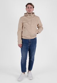 Giacca capovolta beige chiaro con zip frontale e tasche laterali, indossata sopra jeans blu e sneakers bianche. Materiale morbido e leggero.