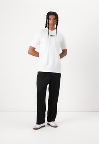 Hvit bomullst-skjorte med svart "adidas" logo, sammen med svarte bukser og hvite joggesko med brune detaljer. Slim fit, glatt tekstur.