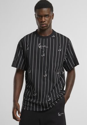 Fiatal férfi fekete, rövid ujjú, fehér függőleges csíkos ingben, kurzív betűs felirattal, fekete nadrággal, amelynek zsebére hasonló szöveg van írva.