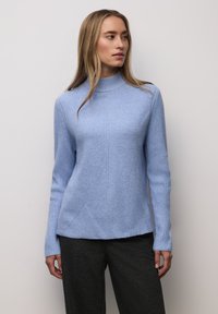 Pitkähiuksinen vaalea nainen pukeutuneena vaaleansiniseen ribbineuleiseen poolopaitaan ja mustiin teksturoituihin housuihin, seisoo yksinkertaista valkoista taustaa vasten.