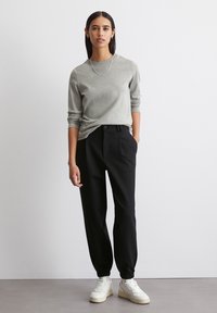 Grå langærmet skjorte parret med sorte joggerbukser, der har elastiske manchetter og lommer, båret med hvide sneakers foran en neutral baggrund.