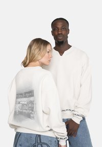 Două persoane care poartă hanorace albe cu manșete decorative; una se află cu fața la cameră, cealaltă arată un grafic alb-negru pe spate.