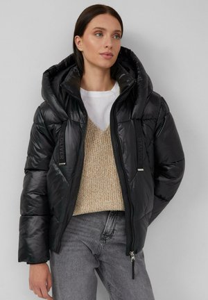 Zwarte pufferjas met een grote capuchon, glanzende textuur, ritssluiting en logo-accenten. Draag het over een beige gebreide trui en grijze jeans.