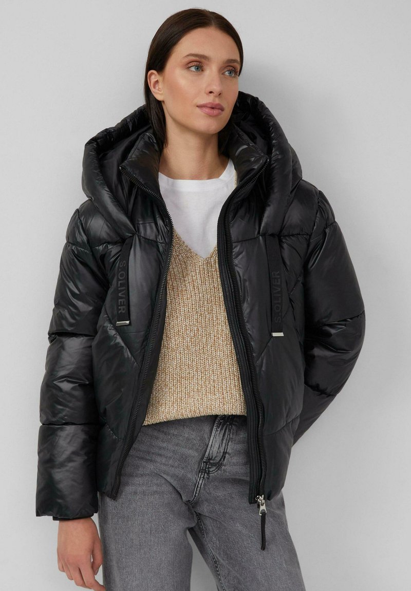 Zwarte pufferjas met een grote capuchon, glanzende textuur, ritssluiting en logo-accenten. Draag het over een beige gebreide trui en grijze jeans.