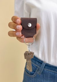 Hnědý kožený držák na klíče s patentním knoflíkem, připevněný na kovovém kroužku na klíče, držící několik stříbrných klíčů. Držen v ruce proti bílé košili.