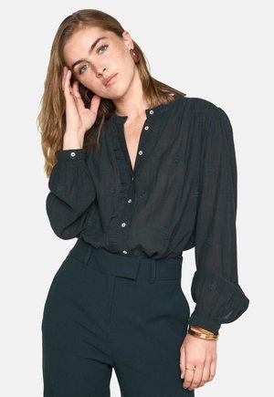 Blusa nera a maniche lunghe con colletto arricciato e chiusura con bottoni. Presenta un delicato ricamo floreale e una vestibilità ampia, abbinata a pantaloni a vita alta.
