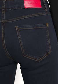 Jean en denim bleu foncé avec une poche arrière dotée de coutures orange contrastantes et d'une étiquette de marque rouge sur la ceinture.