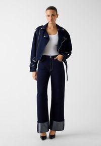 Μπουφάν από τζιν σε ναυτικό μπλε με ασημί εξαρτήματα, εφαρμοστό με ασύμμετρο φερμουάρ, συνδυασμένο με παντελόνι σε ναυτικό μπλε φαρδύ με μανσέτες από τζιν.