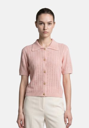 Donna dalla pelle chiara e capelli scuri indossa un cardigan a maglia rosa pallido a maniche corte con bottoni e pantaloni color crema su uno sfondo semplice.