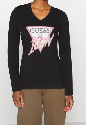 Personne portant un haut noir à manches longues en V avec un logo Guess Icon rose et blanc sur la poitrine et un pantalon marron.