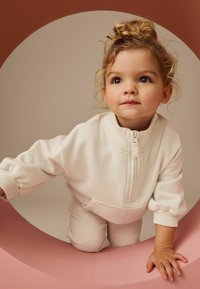 Kermanvärinen, laikukalla fleece-setti, jossa on vetskardefillerio, ribbed-mansetit ja taskut; lapsi poseeraa vaaleanpunaisella pyöreällä rakenteella.