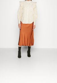 Blusa con maniche a palloncino e motivo floreale, abbinata a una gonna midi in satin arancione e stivali alla caviglia verdi.