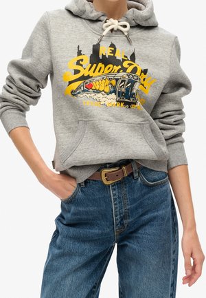 Osoba ubrana w szarą bluzę z kapturem z żółtym napisem "Super Dry", grafiką metra oraz niebieskie dżinsy z brązowym paskiem, ręka włożona do kieszeni.