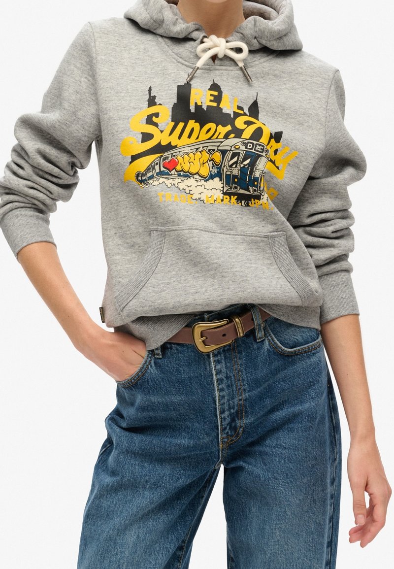 Personne portant un sweat à capuche gris avec texte jaune "Super Dry", illustration de métro, et jean bleu avec une ceinture marron, main dans la poche.