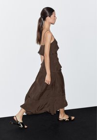 Hnědá, vrstvená, bezrukávová maxi šaty s volány, splývavá látka a jemný lesk. Nosené s černými sandály s minimalistickým designem.