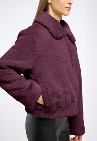 Syntetyczna futrzana kurtka, krótka, w ciemnofioletowym kolorze z szerokim kołnierzem, długimi rękawami i bocznymi kieszeniami. Teksturowana, miękka powierzchnia.