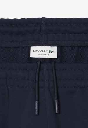 Shorts de sport bleu marine avec taille à cordon, présentant une texture tissée et une étiquette Lacoste. Design à coupe régulière avec des accents cousus.
