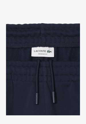 Pantalones cortos deportivos azul marino con cintura ajustable, que presentan una textura tejida y una etiqueta de Lacoste. Diseño de ajuste regular con acentos cosidos.
