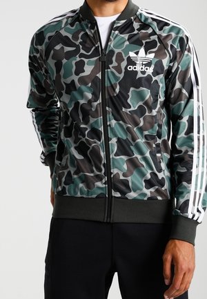Mann trägt eine grün-schwarze Camouflage Adidas Sweatjacke mit Reißverschluss, weißen Streifen an den Ärmeln und Adidas-Logo auf der linken Brust.