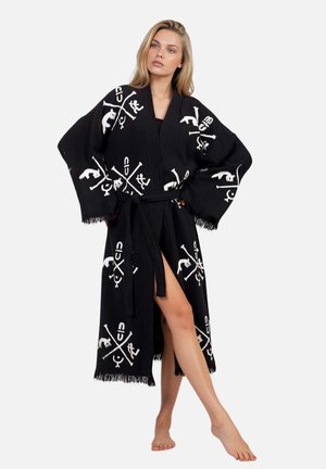 Donna bionda che indossa un accappatoio nero in stile kimono con simboli grafici bianchi, maniche ampie e cintura annodata, in piedi a piedi nudi con una gamba in avanti.