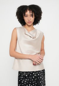 Beige sateng ermeløs topp med drapert hals, kombinert med et svart skjørt pyntet med hvite prikker. Jevn tekstur, moderne silhuett.