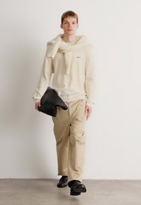 Giovane uomo che cammina all'interno, indossando pantaloni beige, un maglione crema con un capo di maglia sulle spalle, mentre tiene una borsa in pelle nera.