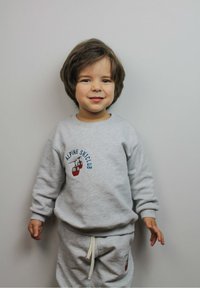 Glunderend jong kind met kort bruin haar, gekleed in een lichtgrijze Alpine Ski Club sweatshirt en bijpassende broek, staande tegen een effen muur.