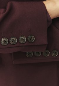 Burgundská vlněná látka s sedmi tmavými, lesklými knoflíky podél manžety, každý se čtyřmi dírkami, a kontrastními švy.