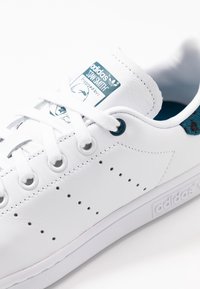 Zapatilla blanca de cuero con detalles perforados, interior turquesa, ojales azules y una etiqueta de logo verde y blanca en la lengüeta.