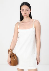 White linen dress with thin straps, square neckline, and side buttons. Brown textured handbag held in one hand.