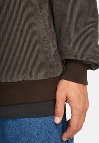 Giacca di velluto a coste marrone con texture a coste, con un polsino marrone scuro. Indossata sopra una camicia scura e jeans azzurri chiari. Mano visibile.