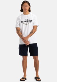 Camiseta blanca de algodón con logo gráfico negro, combinada con shorts cargo azul marino y chanclas negras. El modelo está de pie con los brazos cruzados.