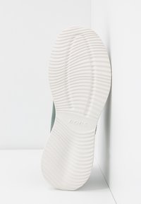 Semelle blanche d'une chaussure avec des rainures horizontales et le mot "BOBS" embossé au centre, incliné contre un mur d'angle blanc.