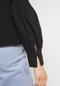 Blusa nera a maniche lunghe puff con polsini abbottonati, abbinata a una gonna o pantaloni in tessuto a coste di colore azzurro chiaro.